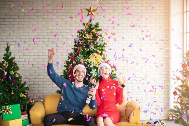 man and woman wearing santa hats sitting on sofa popping a confetti surrounded by christmas trees